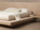 The low platform Petra bed in light leather with ivory linen bedding, a folded throw, and an attached wood nightstand shelf built into the headboard.