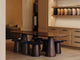 A dining vignette with an oval walnut table on cone-shaped pedestal legs, dark upholstered stools, and wood cabinetry displaying rolled fabric and leather swatches on open shelving.