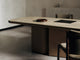 Close-up of the Cota Dining Table with a travertine tabletop and a leather-wrapped plinth base, with a round lacquered bowl and a black tray on top and two of the Sol Dining Chairs in the background.
