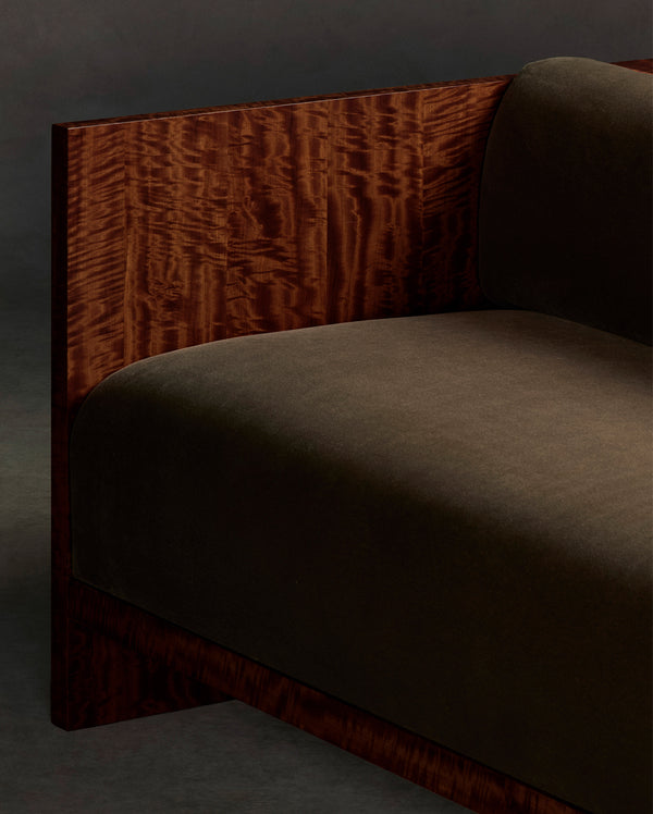 Close-up of a Brasa sofa arm in olive green upholstery with a dark carved wood frame against a dark backdrop.